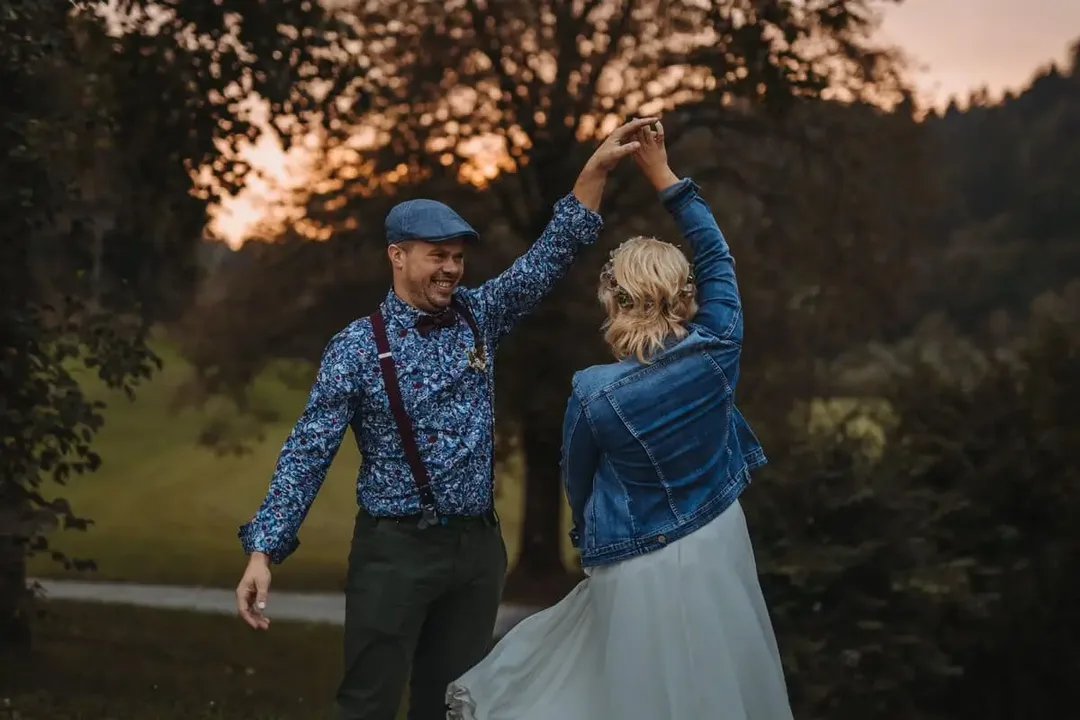 Mladoporočenca zaplešeta na travniku ob sončnem zahodu; ženin v kapi in poslikani srajci zavrti nevesto v jeans jakni.