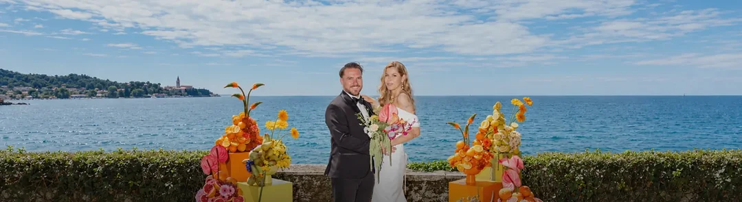 Mladoporočenca na poročnem obredu v Vili Tartini v Strunjanu med dvema kiparskima cvetličnima skupinama s citrusi in cvetjem, za njima odprt pogled na morje in Piran